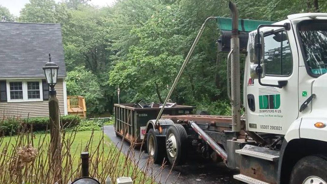 White dump truck with green accents delivering a large dumpster on a residential driveway.