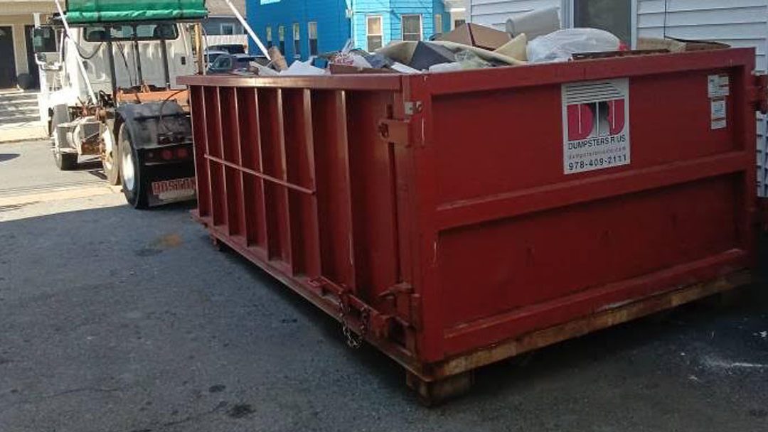 Red metal dumpster filled with trash sits on a wooden pallet in a residential driveway/alley behind a house. Behind it is a small utility vehicle and neighboring homes.