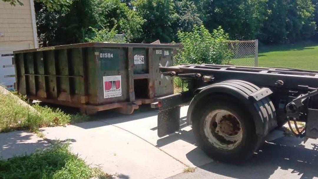 Green dumpster on a trailer attached to a large truck in a residential driveway, ready for disposal work in a sunny yard.