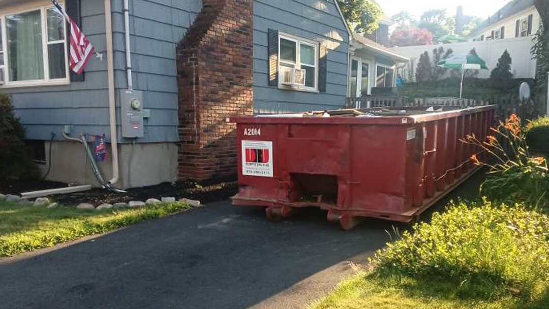 Red dumpster parked in a narrow driveway beside a blue house with an American flag in the yard.
