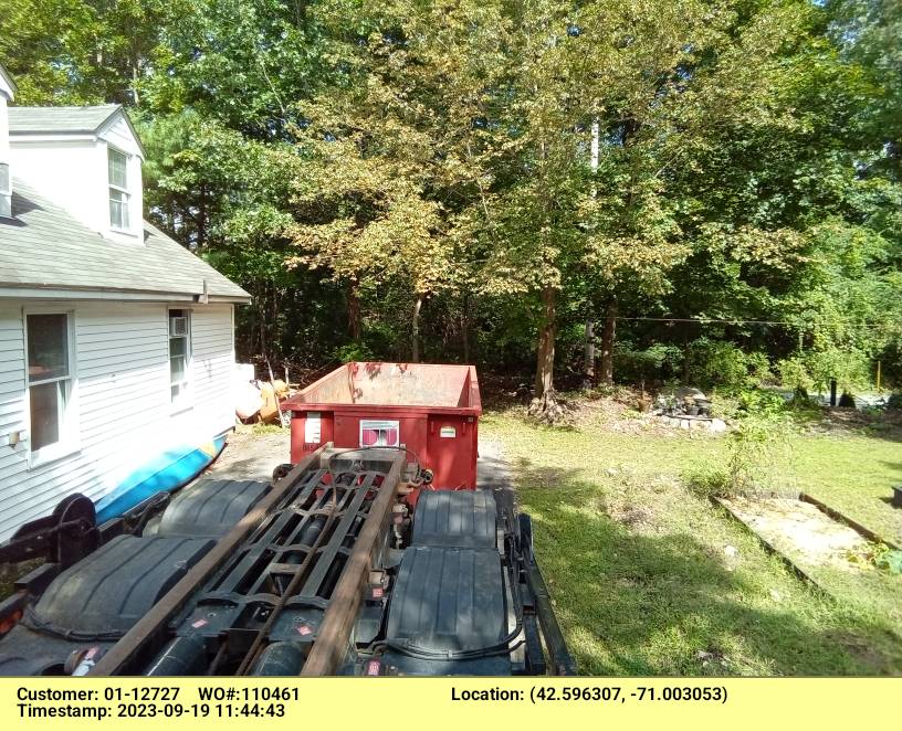 Backyard scene with a white house, a red dumpster, and heavy equipment on a trailer near green trees.