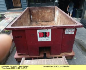 Red industrial dumpster on a wooden pallet in a driveway, with an open rusted interior and a sticker for Dumpster Services R Us visible on the side.