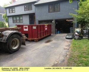 Blue-gray two-story house with an open garage; a large red dumpster on pallets sits in the driveway by a big truck wheel.