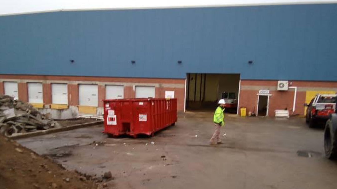 Worker in a high-visibility jacket and hard hat walks past a red dumpster outside a blue warehouse with loading bays.