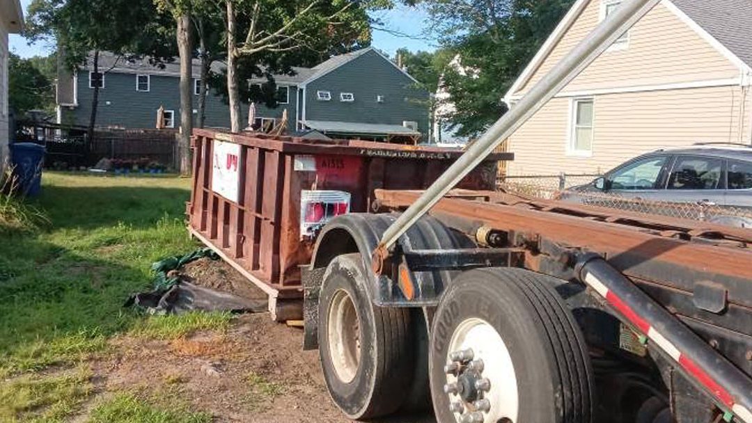 Rusty brown dumpster loaded on a flatbed trailer in a suburban driveway, with a gray car and houses nearby.