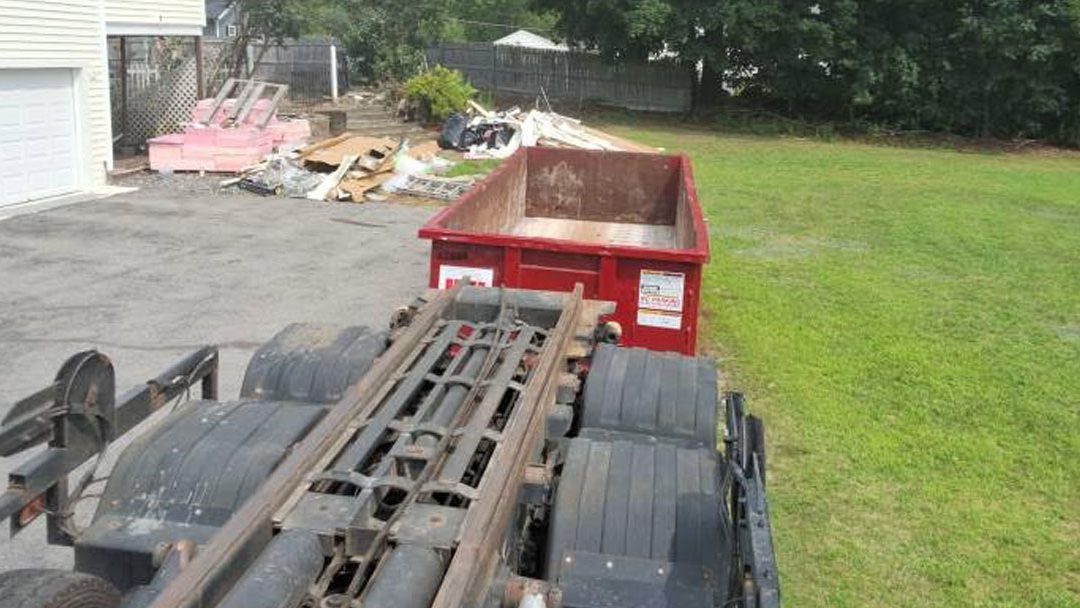 Flatbed trailer in a driveway with a rusted metal frame and a red dumpster filled with debris nearby.