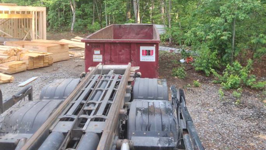 Construction site with stacked lumber, a red dumpster, and a trailer frame on a gravel lot near trees