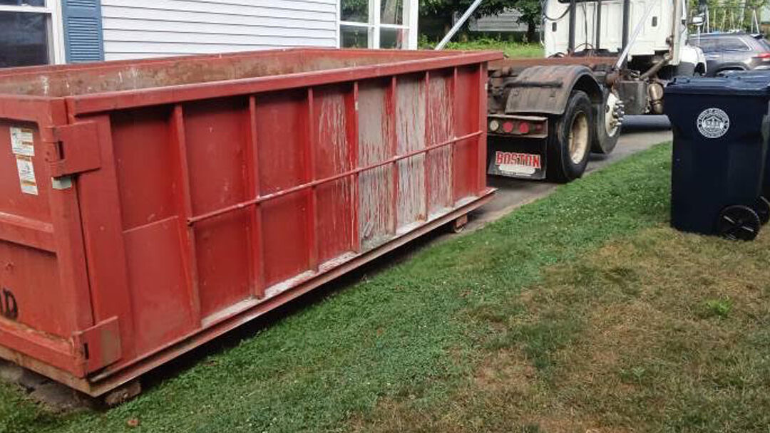 Red industrial dumpster parked on a grassy lawn beside a house, with a garbage truck nearby and two black wheeled trash bins in front of it, curbside.