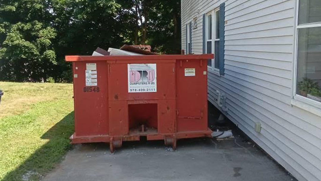 Red dumpster filled with trash sits on a concrete pad beside a light-gray house with blue shutters, outdoors in a yard area.