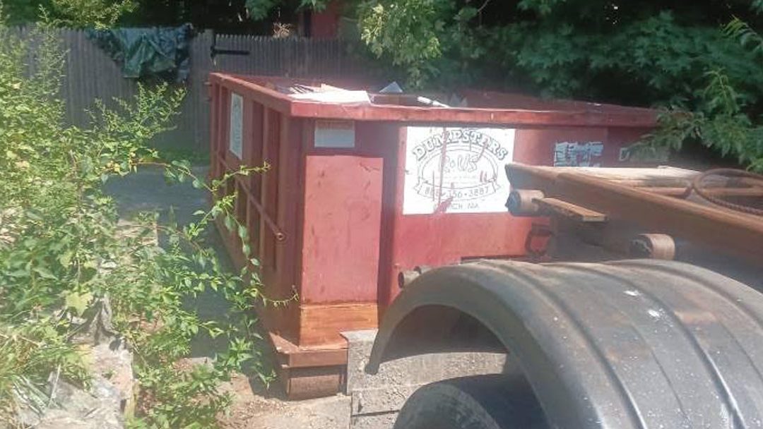 Red dumpster in an overgrown outdoor area with weeds and a trailer hitch nearby.