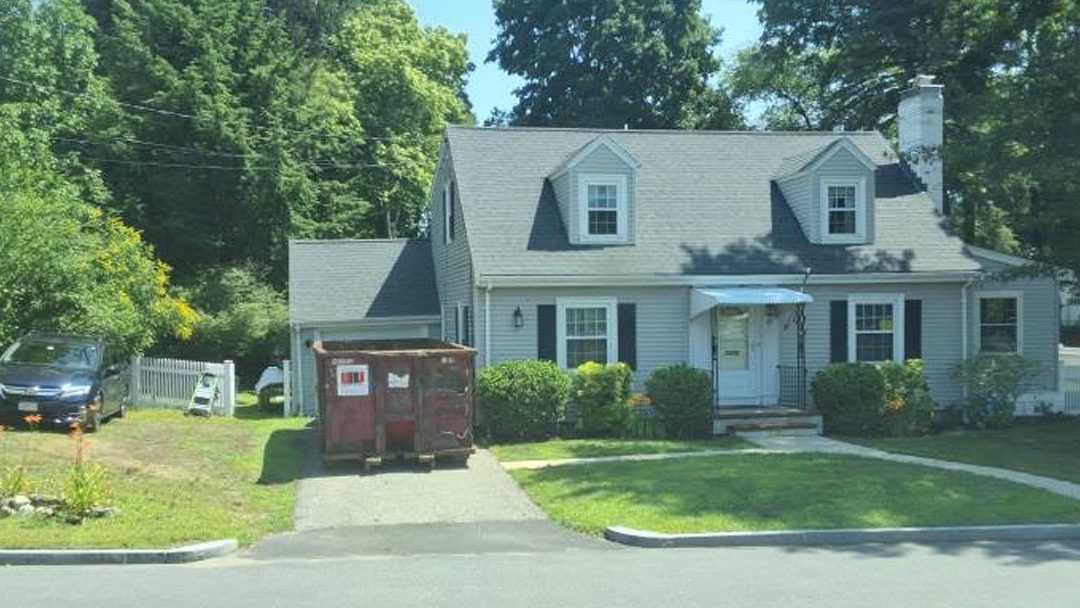 Gray Cape-style house with dormer windows, a blue front door, trimmed hedges, and a red dumpster in the driveway.