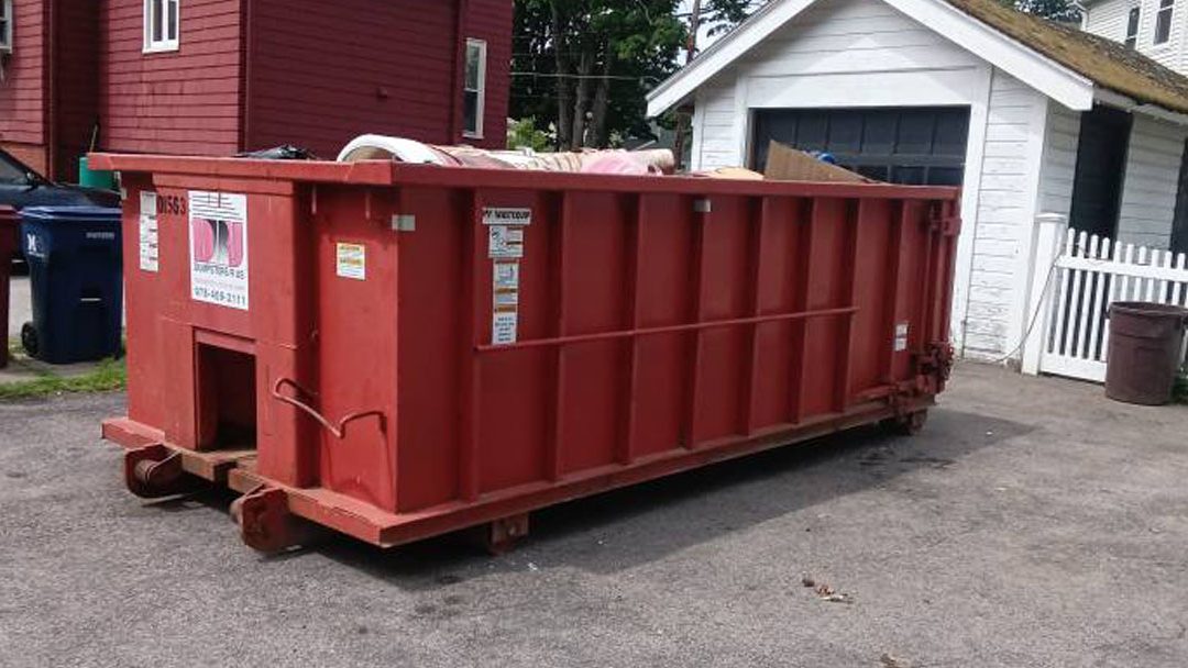 Red dumpster on wheels in a residential driveway near a white fence and garage, with trash bags visible on top.