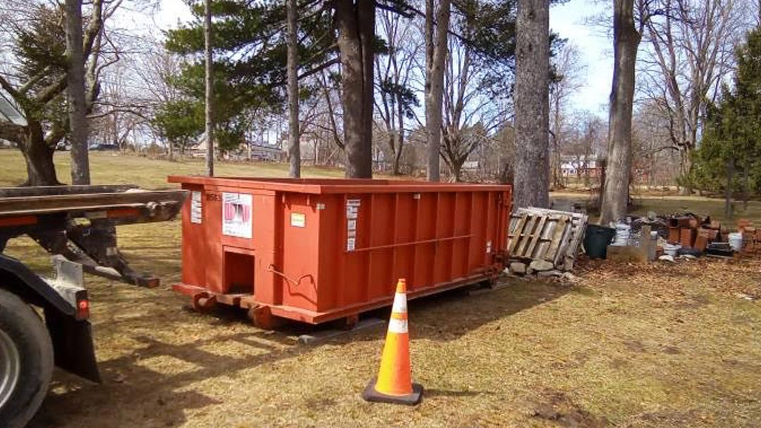 Orange dumpster on wheels in a grassy yard with a traffic cone, pallets, and bricks stacked nearby under trees.